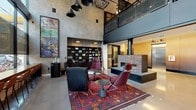 This striking leasing office lobby makes an immediate impression with its soaring double-height ceilings and expansive floor-to-ceiling windows that flood the space with natural light while offering views of the surrounding streetscape. The architectural design blends industrial and modern sensibilities, highlighted by polished concrete floors, exposed structural elements, and a mezzanine level with a sleek metal railing that overlooks the main floor below.

The central lounge area is anchored by a vibrant, patterned kilim area rug and features an eclectic mix of seating, including tufted leather armchairs and mid-century-inspired burgundy chairs, arranged around a minimalist side table. A large-scale, colorful abstract painting commands one wall, adding a bold artistic focal point to the space. Dramatic pendant lighting—a cluster of matte black cone fixtures—descends from the upper volume, complementing the industrial aesthetic.

A built-in shelving unit with organized displays lines one wall, while a decoratively tiled fireplace surround in earthy tones serves as a distinctive architectural feature. Adjacent to the main lobby, a corridor leads past warm wood-paneled walls, elevator bays with brushed steel doors, and a charming mobile beverage cart, offering a welcoming touch. The overall ambiance is sophisticated, creative, and thoughtfully designed to make a lasting first impression.