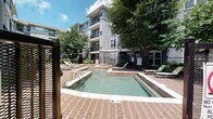 Located at 3949 Lindell Blvd in the heart of St. Louis, MO, this outdoor pool area serves as a central amenity within a multi-story residential community. The space is defined by a clean, rectangular swimming pool with clearly marked depth indicators, accompanied by a shallow wading area — ideal for relaxing in warmer months. The pool deck is finished with classic brick pavers, providing a durable and visually appealing surface that surrounds the water on all sides.

Multiple lounge chairs are positioned around the pool, offering ample space for sunbathing and leisure. Mature trees throughout the courtyard provide natural shade and greenery, contributing to a pleasant outdoor atmosphere. The area is enclosed by a metal security fence with a keyed-access gate, ensuring a private and controlled environment. Signage designates this as a smoke-free and no-glassware zone, reflecting community safety standards.

The pool is set against the backdrop of the multi-story residential building, which features modern gray architecture, private balconies, and large windows. Landscaped garden beds with shrubs and ground cover line the building's exterior, and a concrete walkway connects the pool area to surrounding building entrances, making the space both functional and aesthetically cohesive.