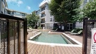 Located at 3949 Lindell Blvd in the heart of St. Louis, MO, this outdoor pool area serves as a central amenity within a multi-story residential community. The space is defined by a clean, rectangular swimming pool with clearly marked depth indicators, accompanied by a shallow wading area — ideal for relaxing in warmer months. The pool deck is finished with classic brick pavers, providing a durable and visually appealing surface that surrounds the water on all sides.

Multiple lounge chairs are positioned around the pool, offering ample space for sunbathing and leisure. Mature trees throughout the courtyard provide natural shade and greenery, contributing to a pleasant outdoor atmosphere. The area is enclosed by a metal security fence with a keyed-access gate, ensuring a private and controlled environment. Signage designates this as a smoke-free and no-glassware zone, reflecting community safety standards.

The pool is set against the backdrop of the multi-story residential building, which features modern gray architecture, private balconies, and large windows. Landscaped garden beds with shrubs and ground cover line the building's exterior, and a concrete walkway connects the pool area to surrounding building entrances, making the space both functional and aesthetically cohesive.