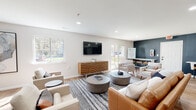 This single-floor community lounge is thoughtfully designed to accommodate a variety of activities, blending social and functional spaces within an open, well-lit layout. Light wood-look flooring runs throughout, complementing the crisp white ceilings fitted with recessed lighting that keeps the space bright and welcoming.

The main lounge area centers around a wall-mounted flat-screen TV positioned above a sculptural wood media console, flanked by a mix of leather and upholstered seating arranged over patterned area rugs. Large windows and a sliding glass door invite abundant natural light and offer views of the surrounding greenery outdoors.

Distinct seating nooks, including high-back tufted chairs arranged for privacy, provide quieter spots for focused work or relaxed conversation. Bold navy and steel-blue accent walls add depth and visual interest throughout, contrasting elegantly with the lighter neutral tones of the furnishings.

A dedicated bar-height counter along one wall serves as a co-working or dining area, equipped with stools. Adjacent to this is a compact kitchenette featuring shaker-style cabinetry, a white countertop, a coffee station, sink, and open shelving adorned with trailing greenery, adding a fresh, organic touch to the space.