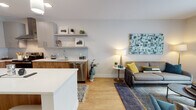 This single-floor residence showcases a thoughtfully designed open-concept layout that seamlessly connects the kitchen, dining, and living areas. The kitchen is a standout feature, offering warm wood cabinetry paired with light gray upper cabinets, a subway tile backsplash, white quartz countertops, and a full suite of stainless steel appliances including a range with a wall-mounted hood. A peninsula island with seating for two is illuminated by pendant lighting, creating a natural gathering point. Floating shelves add both storage and display space above the counters.

The living area is bright and inviting, anchored by light hardwood-style flooring and a sliding glass door that opens to an outdoor space, allowing natural light to fill the room. The bedroom features plush carpeting, a large window with outdoor views, and ample space for furnishings. The bathroom is finished with large-format light tile flooring, a wood-toned vanity, and a tub-shower combination with subway tile surround. Throughout the unit, a neutral color palette, recessed lighting, and modern fixtures create a cohesive, contemporary ambiance.