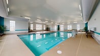 Located at 2015 Wave Crest Dr in Raleigh, NC, this indoor aquatic facility offers a well-appointed, climate-controlled environment for year-round swimming. The centerpiece is a generously sized lap pool with clearly marked lanes, its clear turquoise water reflecting the bright, airy atmosphere of the room. An adjacent hot tub provides a relaxing complement to the pool. The space is designed with high ceilings and an abundance of large windows along the perimeter walls, flooding the interior with natural light and offering views of the surrounding outdoor area. A distinctive teal accent stripe runs along the white walls, adding a cohesive aquatic theme to the décor. The wide pool deck is finished in a neutral tone and is lined with cushioned patio-style seating chairs and small accent tables, creating comfortable areas to relax before or after swimming. A pool lift is available, underscoring the facility's commitment to accessibility. Potted tropical plants are strategically placed throughout the space, enhancing the inviting atmosphere. Flush-mounted overhead lighting supplements natural light for consistent illumination throughout the day. Entry is provided through a glazed door with direct access to the pool deck, making navigation straightforward for all users.