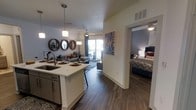 This single-floor residence showcases a thoughtfully designed open-concept layout that seamlessly connects the kitchen, dining, and living areas into one cohesive, flowing space. The kitchen is anchored by a central island with a built-in sink and pendant lighting overhead, complemented by dark cabinetry and modern stainless steel fixtures. Rich, dark hardwood-style flooring extends throughout, adding warmth and continuity to the space. The living area is generously sized, featuring a ceiling fan and large sliding glass doors that open to a private balcony, inviting abundant natural light and outdoor views into the home. Decorative round mirrors and curated wall art lend a contemporary, stylish character to the living space. Adjacent to the main living area, the bedroom offers a comfortable and private retreat, accented by bold wall color and coordinated décor. The open sightlines between rooms create an airy, connected atmosphere throughout the residence. The combination of modern finishes, natural light, and a well-planned layout makes this a highly functional and visually appealing living space suited for everyday comfort and entertaining alike.