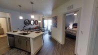 This single-floor residence showcases a thoughtfully designed open-concept layout that seamlessly connects the kitchen, dining, and living areas into one cohesive, flowing space. The kitchen is anchored by a central island with a built-in sink and pendant lighting overhead, complemented by dark cabinetry and modern stainless steel fixtures. Rich, dark hardwood-style flooring extends throughout, adding warmth and continuity to the space. The living area is generously sized, featuring a ceiling fan and large sliding glass doors that open to a private balcony, inviting abundant natural light and outdoor views into the home. Decorative round mirrors and curated wall art lend a contemporary, stylish character to the living space. Adjacent to the main living area, the bedroom offers a comfortable and private retreat, accented by bold wall color and coordinated décor. The open sightlines between rooms create an airy, connected atmosphere throughout the residence. The combination of modern finishes, natural light, and a well-planned layout makes this a highly functional and visually appealing living space suited for everyday comfort and entertaining alike.