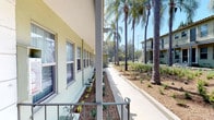 This charming residential property showcases a classic Mediterranean-inspired architectural style, characterized by a stucco exterior finished in a soft sage-green hue, a low-pitched clay tile roofline, and decorative wrought-iron railings that line the covered walkways. The building spans two stories, with the ground floor featuring individual unit entrances accessed directly from a landscaped courtyard pathway.

The exterior grounds have been thoughtfully designed with drought-tolerant landscaping, including a variety of succulents, low shrubs, and native groundcovers set against a clean mulched bed. Majestic palm trees rise prominently throughout the property, providing a tropical ambiance and generous shade across the wide concrete walkways that connect all units.

Each unit along the ground floor benefits from large double-hung windows that allow ample natural light to fill the interiors, while the covered porch overhang provides shade and shelter. The upper-floor units enjoy open-air balconies with wrought-iron railings, offering elevated views of the surrounding greenery and neighboring buildings. The overall atmosphere of the property is bright, airy, and inviting, reflecting a well-kept community setting consistent with a sunny Southern California environment.