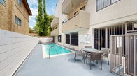 Located at 6210 Woodman Avenue in the Van Nuys neighborhood of Los Angeles, this outdoor pool area offers a well-maintained communal retreat designed for relaxation and recreation. The centerpiece of the space is a generously sized lap pool with clear, blue-green water, tiled edges, and stainless steel entry railings that provide safe and convenient access at both ends.

The surrounding concrete deck is expansive, providing ample room for lounging and movement. A round glass-top table with wicker chairs is positioned along the poolside, creating a comfortable outdoor seating area ideal for gathering. Additional lounge chairs are placed at the far end of the deck for individual relaxation.

The pool area is framed by the multi-story stucco building, which features balconies, security-grated windows, and exterior staircases that contribute to the architectural character of the property. Tall cypress trees and other mature greenery line the perimeter, offering a degree of privacy and a pleasant natural backdrop.

The space benefits from abundant Southern California sunlight, keeping the atmosphere bright and inviting throughout the day. Pool rules are posted prominently on the building exterior, reflecting a well-managed shared amenity. The overall setting is practical, clean, and well-suited for everyday outdoor enjoyment.