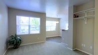This single-floor space offers an open, flowing layout that connects the living and dining areas in a cohesive, functional design. The living area features soft carpeted flooring and is bathed in natural light streaming through a large multi-pane window, which frames views of surrounding greenery. Neutral beige walls and white baseboards throughout create a clean, understated aesthetic that complements a variety of interior styles.

The space transitions smoothly into the dining area, which features hard surface flooring and is illuminated by a pendant light fixture hanging from the ceiling, adding a subtle decorative touch. A second window in the dining area further enhances the brightness of the space. A built-in wall shelf nearby provides convenient display or storage space.

A pass-through window or counter cutout connects the dining area to the adjacent kitchen, promoting an open feel and easy interaction between spaces. The textured ceilings add a modest architectural detail throughout. Overall, the space presents a bright, well-lit, and practical environment with a cohesive neutral palette and comfortable, everyday livability.