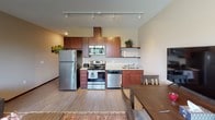 This single-floor unit offers a thoughtfully designed layout that maximizes both comfort and functionality. The open-concept living area serves as the heart of the space, featuring warm hardwood-style flooring, neutral beige walls, and a large multi-pane window arrangement that floods the room with natural light and offers pleasant outdoor views. The living room comfortably accommodates two leather sofas arranged around a central coffee table, all anchored by a decorative area rug that adds warmth to the space.

The kitchen flows seamlessly from the living and dining area, showcasing rich cherry wood cabinetry, granite countertops, and a full suite of stainless steel appliances including a refrigerator, electric range with overhead microwave, and dishwasher. Track lighting overhead ensures excellent illumination across the workspace. A dining table positioned adjacent to the kitchen completes the open entertaining zone.

The bedroom is a calm, private retreat with neutral-toned walls, soft carpeting, and a solid wood door providing privacy. It features ample space for a full-sized bed with an upholstered headboard.

The bathroom is well-finished with tile flooring, a dark wood vanity topped with granite, and a shower-tub combination. Clean, simple design elements throughout give the entire unit a cohesive and comfortable feel.