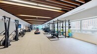 This expansive fitness facility offers a comprehensive, professionally designed workout environment spread across a single, generously proportioned floor. The main gym area is defined by its striking architectural ceiling treatment — rich dark wood slat baffles run the length of the space, complemented by integrated linear lighting that creates a warm yet energizing ambiance. Durable rubber flooring throughout the main floor supports a wide array of equipment, including power racks with weight plates, cable machines, rowing machines, air bikes, treadmills, ellipticals, and a full dumbbell rack with adjustable benches. Plyometric boxes and functional training accessories add versatility to the workout zones. Large windows line the perimeter, flooding the space with natural light and offering sweeping urban views. Full-length mirrors visually expand the space and assist with form. A built-in locker wall provides convenient storage for members. Separate from the main gym floor, a dedicated studio features warm hardwood flooring, floor-to-ceiling mirrors, a mounted display screen, and Pilates reformers — ideal for group classes or focused individual training. Clean white walls and recessed lighting give the studio a calm, focused atmosphere. Together, these two zones create a complete, high-end fitness destination.