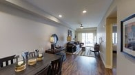This single-floor residence offers a thoughtfully designed open-concept layout that seamlessly connects the dining and living areas into one cohesive, inviting space. Rich, dark hardwood flooring runs throughout, lending warmth and a sense of continuity to the interior. The dining area is anchored by a sturdy dark wood table and chairs, complemented by decorative glass accents that add a refined touch.

The living area is generously proportioned, featuring a comfortable seating arrangement centered on a patterned area rug that defines the space. A ceiling fan with a classic fixture provides both function and a subtle design element overhead. Recessed lighting is installed throughout the ceiling, ensuring even, adjustable illumination across the open floor plan.

Neutral, light-toned walls create a clean and versatile backdrop, allowing natural light to play an important role in the ambiance of the space. A large sliding glass door at the far end of the living area floods the interior with natural light and provides direct access to an outdoor balcony, extending the living space beyond the interior walls. Wall-mounted accent lighting and curated décor details further enhance the polished, well-appointed character of this residence.