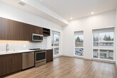 This contemporary single-floor unit offers a thoughtfully designed open-concept layout with clean lines and modern finishes throughout. Light hardwood flooring flows seamlessly across all living areas, complementing the neutral, warm-toned walls and recessed lighting overhead.

The kitchen is a standout feature, showcasing rich dark wood cabinetry with sleek hardware, white quartz countertops, and a large subway tile backsplash. A full suite of stainless steel appliances — including a refrigerator, electric range, built-in microwave, and dishwasher — provides everything needed for functional everyday living. Under-cabinet lighting adds a refined touch to the workspace.

The adjoining living area is bright and open, benefiting from multiple large windows that frame views of the surrounding neighborhood and greenery, filling the space with generous natural light. The elevated ceiling height in this zone enhances the sense of openness and airiness.

A private bedroom offers a quiet retreat, accessed through a paneled door, with hardwood flooring continuing from the main living space. The full bathroom features clean white subway tile surrounding a soaking tub and shower combination, a wood-toned vanity with a white countertop, and hardwood-style flooring, maintaining the cohesive modern aesthetic found throughout the unit.