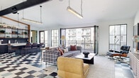 Located at The Overlook in Denver, Colorado, this thoughtfully designed single-floor living space offers a compelling blend of industrial and modern aesthetics. The open-plan layout is anchored by a striking black-and-white checkerboard tile floor that runs throughout, creating a bold visual foundation beneath soaring ceilings with exposed ductwork and chain-hung industrial pendant lighting.

The living area is generously proportioned, accommodating multiple seating arrangements including plaid upholstered sofas, mid-century modern lounge chairs, and a leather accent chair. A wood-slatted credenza with wall-mounted artwork adds warmth and character to one end of the room. Open shelving with warm wood framing provides additional visual depth near the kitchen bar area, which features counter seating and a sleek dark backsplash.

Floor-to-ceiling black-framed windows and a large folding glass door system flood the interior with abundant natural light and provide seamless access to an outdoor balcony. The balcony features open metal grate decking and steel railings, offering elevated views of the surrounding community and courtyard below, including a glimpse of the pool area.

A secondary seating lounge near the entry showcases additional black-framed glass partitions, further enhancing the light-filled, airy ambiance that defines this versatile and well-appointed space.