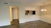 This single-floor space offers a practical and connected layout, highlighted by rich, dark hardwood flooring that runs throughout the main living area, adding warmth and a sense of continuity. The living room is open and unfurnished, providing a blank canvas with clean white walls and white baseboards that keep the space feeling bright and airy. A textured ceiling adds a subtle decorative element overhead.

A defining architectural feature is the pass-through window cut into the wall between the living area and the kitchen, allowing for easy communication and an open feel between the two spaces. The kitchen, visible through the pass-through, features dark cabinetry paired with a mosaic tile backsplash in warm earth tones, offering a stylish contrast to the lighter surroundings. A stainless steel sink and appliances are also visible, contributing to a functional and modern kitchen setup.

A hallway extends from the living room, leading to a doorway with a white-paneled door, suggesting access to additional rooms or storage areas. The overall ambiance is clean, modern, and move-in ready.