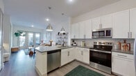 This thoughtfully designed one-bedroom, one-bathroom residence offers a cohesive modern aesthetic across its single floor. The open-concept kitchen and living area serves as the heart of the space, where the kitchen features crisp white shaker cabinetry, a herringbone tile backsplash, sleek gray quartz countertops, stainless steel appliances, and pendant lighting over a peninsula island. Light wood-tone flooring flows throughout the main living areas, complementing the bright, neutral palette. The living room opens to a private balcony through glass French doors, inviting natural light and outdoor views. A clean entryway with a dedicated closet provides a practical transition into the home. The bedroom is a comfortable retreat with large windows overlooking greenery, offering ample natural light. The full bathroom is elegantly appointed with a dual-sink vanity featuring gray cabinetry, white countertops, black-framed mirrors, and a decorative patterned tile floor, accessible directly from the bedroom. A dedicated closet rounds out the layout, providing practical storage. Overall, the space strikes a balance between modern functionality and warm, inviting design.