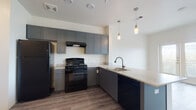 This well-designed single-floor unit offers a clean, modern aesthetic throughout its open and functional layout. The kitchen is a standout feature, showcasing gray shaker-style cabinetry that extends to the upper walls, light countertops, and a full suite of black appliances including a gas range, refrigerator, and dishwasher. A peninsula with an integrated sink and stylish pendant lighting above creates a natural divide between the kitchen and living area, making the space feel connected yet organized. The living area is bright and airy, benefiting from large windows and glass French doors that open directly onto a private balcony with lush outdoor views. A ceiling fan adds comfort to this versatile space, which is finished with durable wood-look flooring that flows consistently throughout. The bedroom is a calm and understated retreat with clean white walls, the same wood-look flooring, and easy access to adjacent spaces. The bathroom is thoughtfully appointed with gray shaker vanity cabinetry, a marble-style countertop, a large mirror with built-in shelving, and a full tub-shower combination with safety grab bars — combining practicality with a polished look. Overall, the unit presents a cohesive, contemporary style with neutral tones and quality finishes.