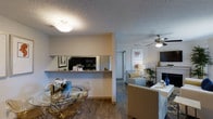 This single-floor residence in Lewisville, Texas, offers a thoughtfully designed open-concept layout that promotes a natural flow between living spaces. The dining area is positioned at the front of the main living zone, featuring wood-look flooring that extends throughout, and is large enough to accommodate a round glass dining table with modern acrylic chairs — a stylish touch that keeps the space feeling open and airy. A half-wall pass-through connects the dining area to the kitchen, which is equipped with dark appliances including a microwave and range. A mirrored backsplash in the kitchen adds depth and reflects light throughout the space. The living room beyond is generously proportioned and anchored by a classic white fireplace, creating a warm and inviting focal point. A ceiling fan with a brushed nickel finish provides both comfort and a contemporary detail to the room. Neutral wall tones and textured ceilings are consistent throughout, lending a cohesive and calm ambiance. Soft, layered lighting from table lamps and flush-mount ceiling fixtures ensures a welcoming atmosphere throughout the day and evening.