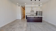 This single-floor space showcases a cohesive modern aesthetic defined by polished concrete floors that extend seamlessly throughout. The open-concept living and kitchen area serves as the heart of the unit, where natural light filters in through floor-to-ceiling black-framed windows, highlighting the clean white walls and industrial-inspired finishes.

The kitchen is a standout feature, equipped with crisp white upper cabinetry contrasted by deep espresso-toned lower cabinets and island base. A white quartz countertop spans the generous kitchen island, which includes bar-height seating brackets, an undermount sink, and ample workspace. Stainless steel appliances—including a gas range, French door refrigerator with ice maker, and a sleek wall-mounted range hood—complement the marble-patterned tile backsplash. Edison-style pendant lights hang above the island, adding warmth to the contemporary palette.

The bathroom continues the minimalist theme with dark cabinetry, a white quartz vanity top, matte black hardware accents, and a glass-enclosed shower. A dedicated closet area features wrap-around hanging rods, offering practical storage. Recessed lighting throughout ensures consistent, even illumination across all spaces.