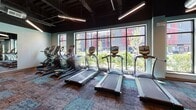 Located at 21 East St in North Attleboro, MA, this fitness center offers a well-equipped, single-floor facility designed to support a full range of workout needs. The space features an open, industrial-inspired aesthetic with exposed black ductwork and structural elements overhead, complemented by sleek linear pendant lighting that illuminates the floor below.

The main workout area is expansive, with textured multi-toned carpet tiles underfoot and soft blue-gray walls that create a clean, energizing atmosphere. A dedicated cardio zone lines the perimeter near floor-to-ceiling grid-pane windows, which flood the space with natural light and provide views of the surrounding outdoor environment. This section houses a row of True-brand treadmills and elliptical machines. Mounted flat-screen TVs are positioned throughout for entertainment during workouts.

The strength training area features a variety of resistance machines, a free weights rack stocked with dumbbells, adjustable benches, and additional cable and plate-loaded equipment arranged to allow for comfortable movement between stations. Mirrored walls in the free weights section enhance the sense of space.

Supporting amenities include a restroom corridor finished with light neutral tile flooring and pale blue walls, complete with a wall-mounted drinking fountain. The overall design balances functionality with a modern, polished aesthetic suited for daily fitness use.