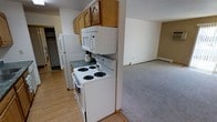 This single-floor residence offers a practical and functional layout designed to maximize everyday living. The kitchen is equipped with white appliances, including a coil-burner electric range, over-the-range microwave, and a full-size refrigerator. Warm-toned wood upper cabinetry provides ample storage, while a dark laminate countertop adds contrast to the bright, light-colored walls. The kitchen features easy-to-maintain hard surface flooring, adding durability and a clean aesthetic to the cooking area. Adjacent to the kitchen, the living space transitions to soft wall-to-wall carpeting, creating a comfortable and inviting atmosphere. The open sightline between the kitchen and living area enhances the sense of space, allowing natural flow between the two zones. The neutral color palette throughout — from the beige walls to the light trim and baseboards — contributes to a cohesive and understated interior. A closet, visible near the kitchen, offers additional storage convenience. The overall design is straightforward and practical, catering to those who value function and ease of maintenance in their daily living environment.