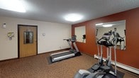 This single-room fitness center offers a practical and well-equipped space for exercise. The room features a distinctive terracotta-red accent wall that adds a bold visual contrast to the surrounding neutral beige walls, creating an energetic atmosphere conducive to working out. Carpet flooring in a warm brown tone runs throughout the space, providing a cushioned surface underfoot.

The cardio section includes a treadmill and an elliptical trainer, both positioned to take advantage of a large wall-mounted mirror that visually expands the room and allows users to monitor their form during workouts. A multi-station cable weight machine and an accompanying bench round out the strength training offerings, making this a versatile space for a full-body workout.

Natural light filters in through windows fitted with vertical blinds, while overhead flush-mount light fixtures ensure bright, even illumination throughout the room. Baseboard heating units along the walls maintain comfort year-round. The room is accessed through a solid wood-framed door with a frosted glass panel, and a paper towel dispenser and hand sanitizer station near the entrance promote cleanliness. A textured ceiling adds a subtle architectural detail that ties the space together. The layout is efficient, with equipment arranged to maximize usability within the available footprint.