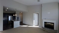 This single-floor space showcases a thoughtfully designed open-concept layout that seamlessly connects the kitchen and living areas. The living room is anchored by a charming fireplace with stacked stone detailing and a classic white mantel, adding warmth and character to the space. Neutral-toned walls and light carpeting create a clean, welcoming ambiance throughout.

The kitchen features dark cabinetry contrasted by a light stone countertop, complemented by stylish pendant lighting that hangs above the kitchen island or peninsula. A sink is integrated into the counter space, and the overall design is functional yet aesthetically refined. The kitchen area transitions smoothly into the adjoining spaces, with recessed lighting and crown molding adding architectural detail overhead.

The open layout benefits from generous natural light streaming through doorways and windows, casting bright patterns across the flooring. A white interior door leads to an additional room or hallway, extending the functional flow of the home. Ceiling fans and HVAC vents are thoughtfully positioned to ensure year-round comfort. The combination of dark wood-toned accents, soft neutral walls, and mixed flooring materials — carpet in the living area and hard flooring in the kitchen — gives this space a balanced, move-in-ready appeal.