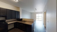 This single-floor unit offers a practical and modern layout designed for comfortable everyday living. The kitchen is a standout feature, showcasing dark espresso-toned cabinetry extending to the upper walls, light-colored countertops, and a full suite of black appliances including a refrigerator, range, dishwasher, and microwave. An L-shaped counter configuration maximizes workspace, while a peninsula with a breakfast bar overhang connects seamlessly to the adjacent living and dining area. Dark hardwood-style flooring runs throughout the main living spaces, adding warmth and visual continuity. The open living area is bright and spacious, benefiting from a large window that invites natural light and offers outdoor views. A carpeted bedroom provides a quieter, softer retreat, complete with its own window and a dedicated HVAC access panel. The bathroom is equipped with an accessibility-friendly design, featuring grab bars along the combination tub and shower, a wall-mounted vanity with a wide mirror, and soft overhead lighting. Neutral wall tones in soft gray and warm beige carry throughout the unit, creating a clean, cohesive aesthetic. The overall layout is efficient and well-organized, making effective use of the available space across each area.