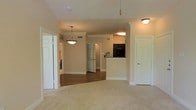 This single-floor residence is designed with an open-concept layout that promotes a smooth flow between the main living areas. The living space features plush carpeting in a warm neutral tone, while the adjacent dining and kitchen areas transition to hardwood-style flooring, creating a subtle but effective visual distinction between zones. Crown molding runs along the ceilings throughout, adding a classic architectural touch that elevates the overall finish of the space.

A peninsula-style kitchen counter defines the boundary between the kitchen and the common areas, offering a functional workspace while maintaining the open feel. Black appliances are visible within the kitchen, contrasting cleanly with the light-colored cabinetry and walls. Overhead lighting includes a stylish pendant fixture that anchors the dining area, while flush-mount ceiling lights provide consistent illumination throughout.

White paneled doors with traditional detailing are found throughout the unit, contributing to a cohesive, classic aesthetic. Neutral warm-beige walls provide a versatile backdrop, making the space feel welcoming and adaptable. Visible HVAC vents are integrated into the ceiling, ensuring practical comfort year-round. Overall, the space presents a well-balanced combination of functionality and classic residential design.