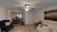 This single-floor residence features an open-concept layout that seamlessly connects the living room and dining area, creating a comfortable and functional flow throughout the space. The living room is carpeted and centers around a classic ceiling fan with integrated lighting, providing both comfort and illumination. A white bifold closet door offers convenient built-in storage, while a front entry door with a wall-mounted fixture adds a welcoming touch.

Adjacent to the living area, the dining space is finished with hardwood-style flooring and is anchored by a traditional dark wood dining table and chairs, set atop an area rug. A pendant light hangs above, adding focused warmth to the area. A pass-through window or open wall connects the dining room to what appears to be a kitchen beyond, where natural light filters in through a window, brightening the space.

The living room is furnished with a white slipcovered sofa accented with orange throw pillows, lending a casual, lived-in charm. Overall, the space presents a warm, functional layout well-suited for everyday living and entertaining.