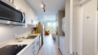 Located on Divine Court in Auburn, Washington, this thoughtfully designed studio offers a smart, functional layout that maximizes every square foot of its compact footprint. The space flows seamlessly from the galley-style kitchen through the living and sleeping area, creating an efficient open-plan arrangement ideal for single occupancy.

The kitchen is outfitted with white shaker cabinetry featuring sleek dark hardware, a stainless steel refrigerator, built-in microwave, dishwasher, electric cooktop, and a double-basin sink with light stone-look countertops. Track lighting overhead ensures the kitchen is well-illuminated. Notably, a stacked in-unit washer and dryer is tucked neatly along the opposing wall, adding significant convenience.

Light wood-tone flooring runs throughout the entire unit, contributing to a bright, cohesive aesthetic. The main living and sleeping area is anchored by a large sliding window that floods the space with natural light and offers pleasant views of surrounding greenery. A built-in closet with sliding doors provides organized storage within the sleeping area.

The bathroom features warm neutral tones, a white vanity with stone countertop, and clean, understated finishes. Overall, the unit presents a modern, efficient living environment with a bright and airy ambiance.