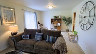 This single-floor space presents an open and functional layout that combines a living area and a home office zone within one cohesive room. The living area is anchored by a large, dark brown corduroy sofa adorned with decorative pillows in coordinating blue, brown, and striped patterns, along with a cozy throw blanket. A flush-mount ceiling light fixture illuminates the space, which features light-colored carpet flooring and neutral beige walls that contribute to a calm, understated ambiance. A large window with vertical blinds on the left wall allows natural light to fill the room.

Toward the back of the space, a wooden hutch desk serves as a dedicated workspace, complete with a rolling office chair and organizational shelving. A potted indoor tree adds a touch of greenery and warmth to the corner nearby. To the right, a partial view into an adjacent area reveals what appears to be a kitchen or dining nook, while a small table with chairs is visible in the background, suggesting additional functional space. Overall, the room feels practical, welcoming, and well-utilized.
