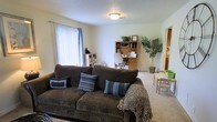 This single-floor space presents an open and functional layout that combines a living area and a home office zone within one cohesive room. The living area is anchored by a large, dark brown corduroy sofa adorned with decorative pillows in coordinating blue, brown, and striped patterns, along with a cozy throw blanket. A flush-mount ceiling light fixture illuminates the space, which features light-colored carpet flooring and neutral beige walls that contribute to a calm, understated ambiance. A large window with vertical blinds on the left wall allows natural light to fill the room.

Toward the back of the space, a wooden hutch desk serves as a dedicated workspace, complete with a rolling office chair and organizational shelving. A potted indoor tree adds a touch of greenery and warmth to the corner nearby. To the right, a partial view into an adjacent area reveals what appears to be a kitchen or dining nook, while a small table with chairs is visible in the background, suggesting additional functional space. Overall, the room feels practical, welcoming, and well-utilized.
