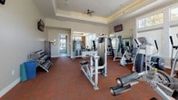 This fitness center is a comprehensively outfitted space designed to support a full range of workout routines. The room features a tray ceiling with crown molding and a ceiling fan, lending the space an elevated, polished aesthetic that sets it apart from a standard gym environment. Recessed lighting complements the abundant natural light that floods in through large, multi-pane windows along one wall, creating a bright and energizing atmosphere.

The flooring throughout is a durable, rich red-toned rubber tile, well-suited for heavy equipment and high-traffic use. A wide array of strength-training machines is distributed across the space, including leg extension and curl machines, a cable crossover system, a multi-functional press machine, a squat rack with weight plates, and a dumbbell rack. Cardio equipment, including a treadmill and elliptical, is positioned near the windowed wall to take advantage of the natural light and outdoor views.

Full-length mirrors along one wall enhance the sense of openness and allow users to monitor their form. Wall-mounted flat-screen televisions are installed in multiple locations for entertainment. A set of double emergency-exit doors on one side provides direct outdoor access, adding convenience and ventilation options.