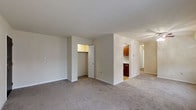 This single-floor unit offers a straightforward, functional layout with a neutral color palette throughout. The main living area is finished with wall-to-wall carpeting in a light beige tone, providing a clean and cohesive look. White walls and a standard flat ceiling keep the space feeling bright and open.

A built-in closet is accessible from the living area, featuring a white panel door and an illuminated interior, offering convenient storage. A partial wall divides the living space from the kitchen area, which is visible in the background. The kitchen features warm, medium-toned wood cabinetry and appears to transition to hard flooring, contrasting with the carpeted living zones.

A hallway visible to the right of the kitchen suggests additional rooms beyond the main living area. The overall design is understated and practical, with simple finishes suited to everyday living. Natural light filters into the space, contributing to its clean, neutral ambiance. The layout prioritizes usable open floor space, making it adaptable for a variety of furniture arrangements and lifestyle needs.