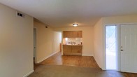 This single-floor space offers a practical and functional layout designed for comfortable everyday living. The main living area is open and airy, featuring warm-toned carpeting that transitions seamlessly into hard surface flooring as it approaches the kitchen. Neutral, light-colored walls and white baseboards create a clean, understated aesthetic throughout.

The kitchen is positioned at the far end of the living space and is defined by a half-wall pass-through that maintains an open connection between the two areas. Warm wood-toned cabinetry spans both the upper and lower sections of the kitchen, providing generous storage. A window above the kitchen sink allows natural light to filter into the cooking area. The kitchen is equipped with standard appliances and counter space suited for daily meal preparation.

A textured popcorn ceiling runs throughout the space, and ceiling-mounted light fixtures provide consistent overhead illumination. A doorway on the left side of the living area leads to additional rooms, while a bright window on the right side of the space introduces natural light and contributes to the overall sense of openness. The unit's straightforward, single-level design makes it an accessible and easy-to-navigate space.