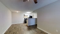 This single-floor space showcases an open-concept layout that integrates the living area and kitchen into one cohesive environment. The flooring throughout is a rich wood-look laminate in a warm, mid-toned finish, lending a sense of continuity and warmth across the space. Neutral gray walls complement the clean white baseboards, creating a modern yet understated aesthetic.

The kitchen is defined by a peninsula-style breakfast bar that separates it from the living area while maintaining the open feel. White upper cabinetry provides ample storage, and the countertops feature a dark granite-style surface. Black appliances, including a range with an overhead ventilation hood and a refrigerator, offer a bold contrast to the lighter cabinetry. A flush-mounted light fixture illuminates the kitchen area, and a visible HVAC vent ensures efficient climate control.

The living area benefits from a ceiling fan with a light kit, supporting both air circulation and ambient lighting. A textured ceiling adds a subtle dimensional detail overhead. The overall ambiance is clean, functional, and move-in ready, with a cohesive modern design throughout.