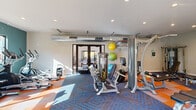 Located at 4501 E Grant Rd in Tucson, Arizona, this fitness center is a fully outfitted exercise facility designed to support a comprehensive workout routine. The single-floor space is thoughtfully arranged to maximize functionality, featuring a diverse selection of strength training machines, including cable and resistance equipment, alongside cardio machines such as treadmills, ellipticals, and stationary bikes. A dedicated dumbbell rack provides a range of free weights, while stability balls in multiple sizes and battle ropes support functional and cross-training exercises. A wall-mounted stretch guide chart offers additional guidance for flexibility training. The flooring consists of a bold geometric-patterned carpet in blue and orange tones, adding a vibrant, energetic aesthetic to the environment. Exposed industrial-style ductwork runs along the ceiling, contributing to the room's modern, utilitarian character, while recessed lighting ensures the space is well-lit throughout. A built-in water fountain station provides convenient hydration access. Double glass doors on two sides of the room allow natural light to filter in and offer views of the exterior surroundings, creating an open and welcoming atmosphere. The space is clean, organized, and well-maintained, making it a practical and inviting fitness facility.