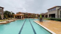 This expansive outdoor amenity space is defined by its resort-style pool area, showcasing a generously sized lap pool with clearly marked lanes and an adjoining spa. The pool deck is finished in warm-toned stamped concrete and brick pavers, creating an inviting atmosphere for relaxation and recreation. Throughout the space, lounge chairs and bar-height seating with market umbrellas are thoughtfully arranged to provide both sun and shade options for residents.

The surrounding landscape is meticulously maintained, featuring lush greenery, manicured shrubs, ornamental trees, and well-kept turf areas that soften the hardscape and add natural beauty to the environment. A covered outdoor pavilion with a distinctive copper-toned roof provides an additional shaded gathering area, complete with seating for socializing.

The community buildings that frame the pool area present a blend of architectural styles, including classic brick facades with pitched rooflines as well as more contemporary stone and stucco exteriors with flat and gabled rooflines. Balconies and large windows overlook the outdoor space, integrating the interior living areas with the amenity below. Decorative iron fencing encloses the pool area, ensuring privacy and security while maintaining an open, airy feel throughout this well-appointed outdoor retreat.