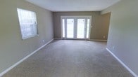 This single-floor space centers around a generously proportioned main living area finished with wall-to-wall plush carpeting in a warm neutral tone, providing a comfortable and inviting foundation. The walls are painted in a soft gray-taupe hue, complemented by crisp white baseboards and trim that add a clean, polished contrast throughout the room. A textured ceiling adds subtle visual interest overhead.

Natural light flows freely into the space through a set of white-framed French doors positioned along the back wall, each flanked by sidelights that frame outdoor views of surrounding greenery. An additional window on the adjacent wall further brightens the interior, ensuring the room feels open and airy throughout the day. The French doors serve as a functional focal point, connecting the interior to the outdoors while enhancing the overall sense of space.

The layout is open and unobstructed, offering flexible arrangement potential to suit a variety of uses. White electrical outlets are neatly positioned along the walls, and the clean lines of the trim and door framing contribute to a tidy, well-maintained aesthetic. Overall, the space presents a neutral, move-in-ready environment with an understated, functional character.