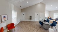 Located at 2295 Pineview Ln in Gastonia, NC, this thoughtfully designed single-floor commercial space blends modern aesthetics with practical functionality. The main area immediately impresses with its soaring vaulted ceilings and a striking gold sputnik chandelier that anchors the room with a contemporary flair. Large glass-panel French doors flood the space with natural light, while a wall-mounted flat-screen television and a dedicated workstation with blue velvet seating make it well-suited for meetings or client-facing operations. Throughout the space, a distinctive herringbone-patterned hardwood-style flooring adds a refined, design-forward touch that ties each area together cohesively. A separate breakroom or lounge area features a marble-top round dining table with coordinating blue chairs and a colorful area rug, providing a comfortable space for informal gatherings. The kitchenette is outfitted with crisp white shaker-style cabinetry, a stainless steel refrigerator, a microwave, and a sink with ample counter space. A hallway area includes two dedicated restrooms and a wall-mounted water fountain, ensuring full convenience for staff and visitors alike. The neutral warm-toned walls and recessed lighting throughout create a clean, professional ambiance that is both welcoming and versatile.