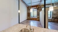 This loft apartment in Charlotte's 28205 zip code blends industrial character with modern upgrades. Soaring ceilings with exposed ductwork and structural beams define the open living area, which features polished concrete floors and a striking exposed brick accent wall with large windows that invite natural light. The kitchen is well-appointed with dark cabinetry, granite countertops, stainless steel appliances, and a peninsula with an undermount sink and pendant lighting overhead. The bathroom includes a full tub-shower combo with clean, bright finishes. The overall ambiance is urban and contemporary.