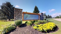 The Tanglewood community clubhouse is a welcoming, single-floor gathering space that makes a strong first impression with its beautifully landscaped entrance sign, framed by stone pillars and vibrant seasonal plantings. Inside, the space is defined by dramatic vaulted ceilings with exposed beam detailing and multiple ceiling fans, creating an airy, open atmosphere throughout. The main lounge area is generously sized, offering plush sectional and accent seating arranged for comfortable socializing. Large windows with vertical blinds line the walls, flooding the interior with natural light and providing views of the surrounding grounds. A standout feature of the lounge is the full-height brick fireplace with a mounted flat-screen television, anchoring one end of the room and adding warmth and character. Built-in window seats provide additional casual seating along the perimeter. Adjacent to the lounge, a dedicated kitchenette and bar area features a tiled countertop bar with barstool seating, a refrigerator, microwave, and prep appliances — ideal for community events and gatherings. A granite-topped high table with stools near the entry serves as a welcoming reception or registration point. Tile flooring in the kitchen zone transitions to carpet in the lounge, and large sliding glass doors open the space to the outdoors, enhancing the sense of connectivity with the community's natural surroundings.