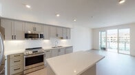 This contemporary single-floor unit is thoughtfully designed with a clean, modern aesthetic and an open, airy layout. The kitchen serves as the centerpiece of the living space, showcasing soft grey shaker-style cabinetry with matte black hardware, a crisp white subway tile backsplash, and light-toned quartz countertops. A full suite of stainless steel appliances — including a refrigerator, range, built-in microwave, and dishwasher — provides a fully equipped cooking environment. A generous kitchen island with a matching quartz surface offers additional prep and gathering space, flowing seamlessly into the open living area. Recessed lighting illuminates the kitchen, while large windows and a glass door admit ample natural light into the living zone, with views of the surrounding neighborhood. Light wood-toned flooring runs consistently throughout the unit, reinforcing a cohesive, modern feel. The bedroom is a serene, well-proportioned space featuring a built-in closet with double hanging rods and a window that brings in natural light. The full bathroom is finished in neutral tones with light wood flooring, a vanity with matching cabinetry, and a combination tub and shower with a curved glass enclosure. Overall, the unit presents a bright, clean, and functional living environment with quality finishes throughout.