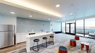 Located at 401 A St in Wilmington, Delaware, this single-floor communal space combines a fully equipped kitchen with an open living area, offering a well-designed environment for gathering and relaxation. The kitchen is a standout feature, appointed with sleek white cabinetry, quartz countertops, a stainless steel refrigerator, an under-counter appliance suite, and a statement backsplash of elongated hexagonal light-blue tiles. A white kitchen island with seating for three provides a casual dining and social focal point, illuminated by a trio of pendant lights suspended from a softly lit tray ceiling. The living area flows naturally from the kitchen, anchored by a blue-toned, abstract-patterned carpet that runs throughout the space. A curated gallery wall displaying an eclectic collection of framed artwork adds a creative, inviting character to the lounge. Comfortable seating, including a tufted linen sofa and rounded wooden ottoman stools, encourages informal use of the space. Floor-to-ceiling windows and glass double doors open directly onto an outdoor terrace, flooding the interior with natural light and offering panoramic views of the surrounding Wilmington skyline and waterfront. Recessed lighting throughout ensures consistent, even illumination across the entire space.