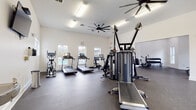 Located at 9015 Kings Grant Dr in Charlotte, NC, this well-equipped fitness center offers a comprehensive workout facility spread across a single floor with two distinct exercise zones connected by an open passage. The space features bright white walls, rubber-speckled flooring designed for high-impact activity, and generous ceiling heights that create an open, airy atmosphere. Large ceiling fans are mounted throughout, ensuring effective air circulation during workouts.

The primary exercise area is lined with windows overlooking an outdoor pool area, allowing ample natural light to fill the space. This zone is outfitted with multiple treadmills with individual screens, an elliptical trainer, a stationary spin bike, and a multi-station cable machine for versatile strength training. A secondary strength-focused room opens directly off the main area and houses a power rack with a bench and barbell, an air resistance bike, and a full rack of dumbbells in varying weights, along with battle ropes for functional training. Recessed lighting and additional ceiling fans keep this area bright and comfortable. Together, the two rooms provide a well-rounded environment suited for both cardiovascular conditioning and resistance training, making this facility a practical and fully functional amenity for the Kings Grant community.