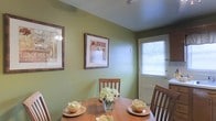 This single-floor space features a charming dining area defined by warm sage green walls that create an inviting, relaxed atmosphere. The round wooden dining table serves as the room's centerpiece, surrounded by matching wooden chairs with slatted backs. The table is styled with decorative bowl centerpieces and a glass vase of white floral arrangements, adding an elegant yet understated touch to the setting.

Two framed art prints with dark wooden frames adorn the green walls, contributing visual interest and a curated aesthetic to the space. Natural light filters into the room through a white-framed door fitted with horizontal blinds, offering a glimpse of greenery outside and enhancing the room's warm, airy feel.

The ceiling features a flush-mounted light fixture that provides overhead illumination, complementing the natural light. The overall ambiance of the dining area is cozy and welcoming, with the combination of earthy green tones, warm wood furnishings, and decorative accents creating a space well-suited for intimate dining and everyday gatherings.