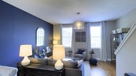 This multi-floor property spans three levels, offering a well-designed living environment with thoughtfully curated interior details. The living room serves as a standout feature of the space, showcasing a deep navy blue accent wall that creates a bold and sophisticated atmosphere. A decorative arched mirror mounted on the accent wall adds architectural charm and visual depth to the room.

Natural light flows generously through multiple large windows dressed with sheer white curtains, brightening the interior and offering views of the surrounding neighborhood. A contemporary geometric pendant light fixture hangs centrally from the ceiling, providing warm ambient illumination that complements the room's layered lighting scheme, which also includes table lamps of varying heights and styles.

The living room is furnished with a substantial sectional sofa in a neutral gray tone, paired with a coordinating armchair and a low-profile area rug that anchors the seating arrangement. Hardwood flooring runs throughout, adding warmth and continuity to the space. Decorative accents, including a wooden wall panel, small sculptures, and coordinated throw pillows, contribute to the cohesive and polished aesthetic of the overall design.