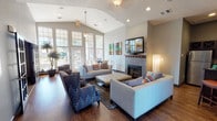 This well-appointed single-floor communal space offers a thoughtfully designed layout that balances comfort with functionality. The main lounge area is anchored by a striking tile-surround fireplace topped with a wooden mantel and a wall-mounted flat-screen television, creating a warm and inviting focal point. Soaring vaulted ceilings amplify the sense of openness, while rich hardwood flooring runs throughout, adding warmth and continuity to the space.

Generous seating arrangements — including neutral sofas, tufted ottomans, and patterned accent chairs in blue — are complemented by colorful contemporary artwork displayed on the walls. Tall, multi-pane windows along one side of the lounge flood the interior with natural light and provide views of the outdoors, enhancing the bright and airy atmosphere.

A dedicated entryway with tile flooring and a wood-and-glass front door provides a welcoming transition into the space. Adjacent to the lounge, a kitchenette or wet bar area features dark cabinetry, bar-height seating with upholstered stools, and pendant lighting overhead.

A separate room, accessible through elegant glass-paned French doors, serves as a business center equipped with a television, work surfaces, and bar-height tables and chairs — ideal for productivity or informal meetings. The overall ambiance is modern yet comfortable, with a cohesive neutral color palette accented by bold, vibrant touches throughout.