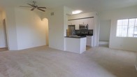 This single-floor residence features an open-concept design that connects the living area to the kitchen, creating a cohesive and functional layout. The spacious living area is finished with light-toned wall-to-wall carpeting, contributing to a clean and neutral aesthetic. A ceiling fan is installed overhead, providing both air circulation and a practical design element.

The kitchen is accessible via an open archway and is defined by a peninsula-style breakfast bar with a dark countertop, offering both additional prep space and a casual dining option. White cabinetry lines the upper and lower sections of the kitchen, providing ample storage, while stainless steel appliances — including a refrigerator, microwave, and range — add a modern touch. A dark tile backsplash offers a subtle contrast to the white cabinetry. The kitchen is finished with tile flooring, distinguishing it from the carpeted living space.

An arched doorway on one side of the living area hints at additional rooms beyond, while the mirrored wall visible in the background adds depth and reflects light throughout the space. Overall, the unit presents a clean, functional, and move-in ready environment.