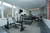 This modern fitness center is designed to offer a comprehensive workout experience in a clean, contemporary setting. The space is thoughtfully laid out across a single level, with distinct zones for both cardio and strength training. A row of treadmills and elliptical machines lines the perimeter, each positioned to take advantage of the large, floor-to-ceiling windows that flood the room with natural light and provide views to the outside. A wall-mounted flat-screen television is conveniently positioned for entertainment during cardio sessions. The strength training area features a multi-function cable machine and weight bench, providing users with versatile resistance training options. Free weights and stability accessories, including an exercise ball, are also available. Underfoot, durable rubber flooring tiles provide safety and support throughout the workout area. Recessed ceiling lighting supplements the natural light, ensuring the space remains bright at all times. A subtly illuminated ceiling detail adds a warm architectural accent. A standout design feature is the large black-and-white photographic mural depicting an urban skyline, which serves as a dramatic focal point and infuses the space with a bold, metropolitan character. The overall ambiance is sleek, functional, and motivating.