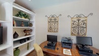 This single-floor space offers a thoughtfully designed layout that balances functionality with classic décor. The business center is a standout feature, equipped with dual computer workstations, a multifunction printer, and a warm wood desk, all set against neutral walls adorned with ornate wrought-iron wall art. A built-in white bookcase provides ample display and storage space, showcasing decorative accents including vases, sculptural pieces, and greenery with teal-painted shelf backs adding a pop of color.

Adjacent to the business area, a spacious built-in entertainment wall unit in white houses a flat-screen television and offers generous shelving for décor. Comfortable upholstered seating and wood-look flooring contribute to a relaxed, inviting atmosphere. A dining or meeting table with upholstered chairs sits near a window with slatted blinds, allowing for natural light.

The open common area is architecturally distinguished by a row of white decorative columns that frame the entry, creating an elegant transition between spaces. Light tile flooring anchors a bright rotunda-style area with tall windows and French doors, while the adjoining lounge area features wood-look flooring, a curved sofa, and a service counter, lending the space a welcoming and communal character throughout.